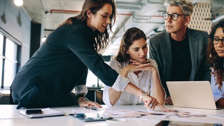 Personen in Businesskleidung stehen rund um einen Besprechungstisch mit Unterlagen, Laptop und Tablets und unterhalten sich, im Hintergrund zeigt sich eine Räumlichkeit im Industrial Style