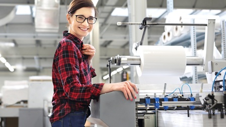 Eine Person mit Brille steht seitlich vor einer großen Maschine. Auf einer Fläche der Maschine liegt ein hoher, weißer Papierstapel. Schräg über dem Stapel ist eine Metallrolle, auf der eine dicke, weiße Rolle ist. Die Person blickt direkt in die Kamera.