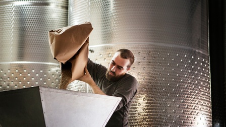 Bärtige Person kippt aus großem Papiersack Getreide in Metalltrichter, im Hintergrund große Metallfässer