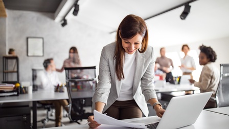 Person mit langen dunklen Haaren und Businesskleidung steht vor einem Bürotisch und blickt dabei auf Unterlagen während sie einen Laptop bedient, im Hintergrund sitzen und stehen weitere Personen rund um Schreibtische in einem Büroraum