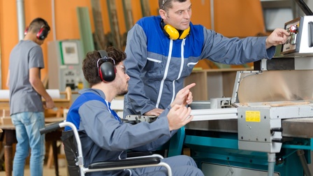 Zwei Personen in Werkhalle an Werkanlagenpaneel: Eine Person bedient Anlage andere Person deutet darauf, beide Personen tragen Schutzbrillen und Gehörschutz, im Hintergrund verschwommen weitere Person