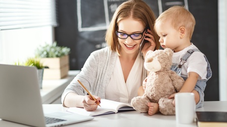 Person mit schulterlangen braunen Haaren und Brille telefoniert und macht sich daneben Notizen in ein Buch während ein Kleinkind daneben auf dem Tisch sitzt und einen Teddybär hält, am Tisch steht auch ein Laptop, eine Tasse sowie Grünpflanzen