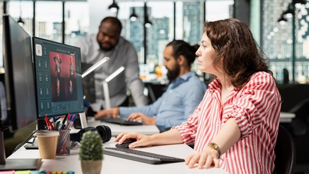 Personen in einem Büro arbeiten an Computern, eine Person sitzt am Schreibtisch und nutzt Maus und Tastatur, zwei weitere Personen stehen und sitzen im Hintergrund.