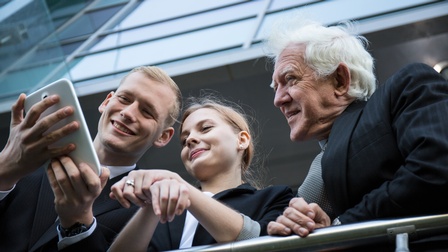 Drei Personen in dunklen Anzügen stehen dicht nebeneinander an einem Geländer und lehnen sich an. Die Person links hält ein Tablet in den Händen. Alle drei schauen in den Bildschirm