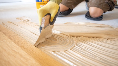 Nahaufnahme der rechten Hand einer Person, die einen Spachtel auf den Boden hält. Am Boden und am Spachtel ist eine weiße, flüssige Masse. Vor der Masse ist ein heller Holzboden. Dahinter sind die Knie einer Person auf einem hellen Boden