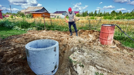 Eine Person schaufelt Sand, er steht neben einer Grube, aus der ein Brunnen ausgehoben wird. Neben ihm ist eine Tonne aus Metall. Im Hintergrund eine Landschaft mit Häusern, es herrscht Sonnenschein. 