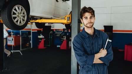 Person in blauem Overall mit Schraubenzieher in der Hand in Werkstatt stehend im Hintergrund Hebetribüne mit Auto