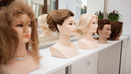Verschiedene Puppenköpfe mit unterschiedlichen Haarfarben und Frisuren sind auf einem Frisiertisch vor einem Spiegel platziert