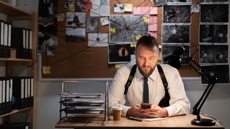 Eine Person sitzt an einem Schreibtisch. In beiden Händen hält sie ein Smartphone, auf das sie blickt. Hinter ihr hängen an einer Pinnwand verschiedene Fotos, die durch rote Fäden miteinander verbunden sind. Auf einem Foto liegt eine Person am Boden