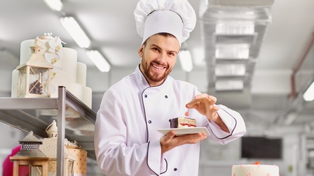Person in weißer Arbeitsbekleidung steht in einer Backstube und hält ein Gebäck in der Hand