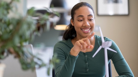 Lächelnde Person sitzt an Schreibtisch und deutet auf Modell eines Windrades