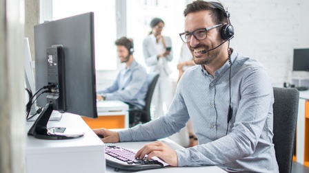 Person mit kurzen dunklen Haaren, Brille und Headset sitzt freudig an einem Schreibtisch und blickt auf einen Monitor, während mit der Tastatur geschrieben wird, im Hintergrund zeigen sich weitere Personen bei Schreibtischen