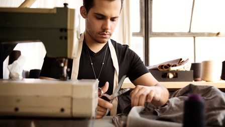 Person sitzt an Tisch und schneidet mit Schere Stoff, im Vordergrund verschwommen im Ausschnitt Nähmaschine, im Hintergrund verschwommen emailliertes Fenster und Tisch mit Zwirnen und Stoffmustern