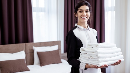 Person mit dunklen, geschlossenen Haaren und schwarz-weißer Arbeitskleidung hält freudig einen Stapel weißer Handtücher in den Händen, im Hintergrund steht ein Bett die Vorhänge bei der Fensterfront sind weiß und in dunklem Lila gehalten