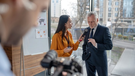 Person hält Mikrofon zu anderer Person, im Vordergrund verschwommene Ansicht einer Person, die Interview mit Kamera aufzeichnet