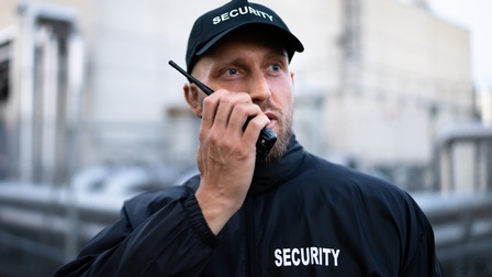Frontale Aufnahme einer Person mit Bart, dunkler Jacke und dunkler Kappe, auf denen jeweils Security steht. Die Person hält sich ein Walkie Talkie vor den Mund. Im Hintergrund ist ein Gebäude