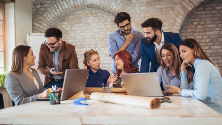 Um einen großen Tisch in einem Büro sitzen fünf Personen. Drei weitere Personen stehen neben dem Tisch. Vor zwei Personen steht jeweils ein Laptop. Die Personen reden in Kleingruppen miteinander und blicken sich an