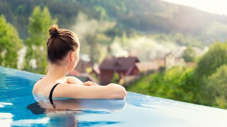 Rückenansicht einer Person, die in einem Pool ist und sich mit den Ellbogen am Beckenrand abstützt. Die Person blickt in die Ferne. Im Hintergrund verschwommen ist eine bergige Landschaft mit Bäumen und Häusern.