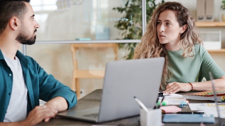 Personen sitzen an einem Schreibtisch und unterhalten sich in einem Büro, ein Laptop und weitere Unterlagen liegen am Tisch
