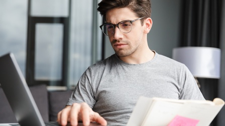 Eine Person mit Brille an einem Tisch. Vor der Person steht ein geöffneter Laptop, den sie mit einer Hand bedient. Rechts daneben liegt ein geöffneter Notizblock mit einem pinkfarbenen Klebezettel. Die Person blickt auf den Bildschirm des Laptops