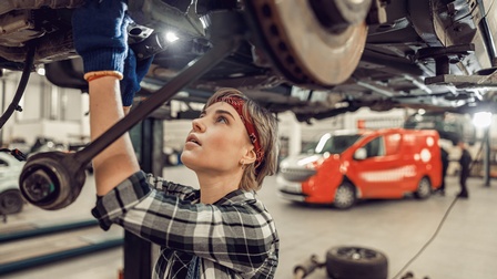 Eine Person mit Handschuhen steht unter einem über ihr fixierten Auto. Die Person hält in der rechten Hand eine Taschenlampe und blickt auf den Unterboden des Autos. Im Hintergrund stehen weitere Autos im Raum