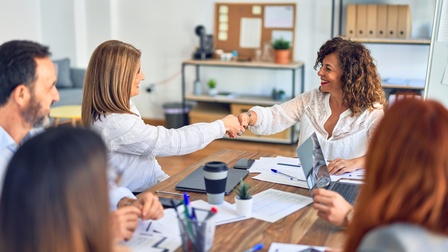 Mehrere Personen um Besprechungstisch sitzend, auf dem Arbeitslunterlagen, Kaffeebecher, und Laptops platziert sind, zwei Personen lächeln einander an und schütteln sich die Hand