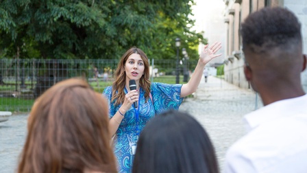 Frontale Aufnahme einer Person mit Mikrofon und Ausweisschild, die im Freien neben einem Gebäude steht und zu einer Gruppe Personen spricht. Hinter der Person ist ein Park