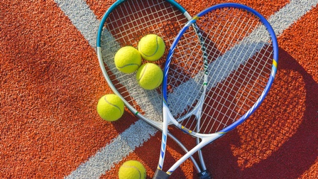 Zwei Tennisschläger sowie fünf gelbe Tennisbälle liegen auf einem roten Untergrund mit weißer Markierung
