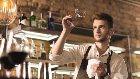 Person mit kurzen braunen Haaren und Bart steht hinter einer Bar und poliert ein Weinglas