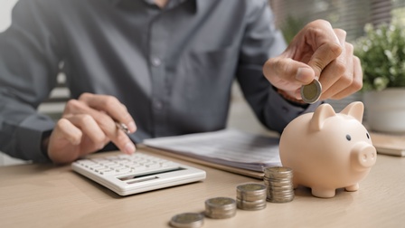 Eine Person tippt mit einer Hand auf einem Taschenrechner und wirft mit der anderen eine 2-Euro-Münze in ein Sparschwein. Auf dem Tisch liegen Münzstapel sowie Zettel mit tabellarischen Auflistungen.