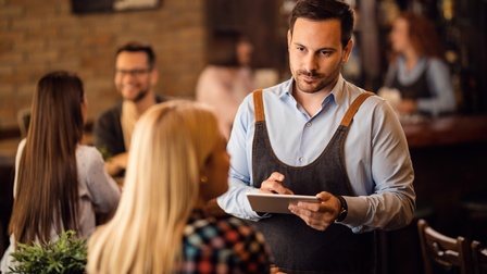 Rückenansicht einer Person, die an einem Tisch in einem Lokal sitzt. Ihr gegenüber steht eine Person mit Schürze, kurzen Haaren und Bart, die ein Tablet hält und zu der sitzenden Person blickt