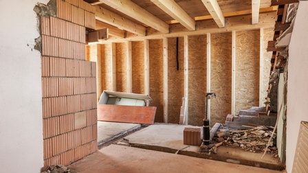 Ein Wohnraum mit Ziegelmauern, Holzwänden und einer Holzdecke. Am Boden ist Beton. In manchen Ecken liegt Schutt