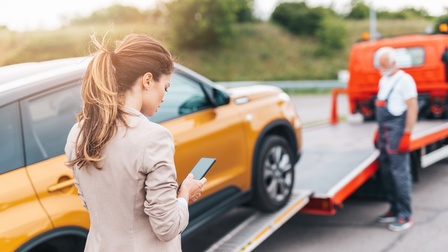 Person mit geschlossenen langen Haaren blickt auf ein Smartphone, während im Hintergrund eine Person in Arbeitskleidung steht und ein Auto abschleppt