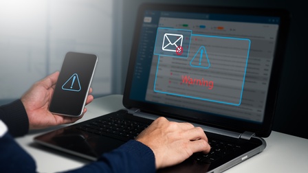 Die Hände einer Person sind auf der Tastatur eines aufgeklappten Laptops. Auf dem Bildschirm ist ein Rufzeichen in einem Dreieck, unter dem Warnung steht. Die Person hält ein Handy, auf dem ebenfalls ein Rufzeichen in einem Dreieck ist