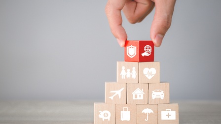 Holzwürfel stehen zu einer Pyramide gestapelt auf einer hölzernen Oberfläche. Auf den Würfeln sind Symbole, zum Beispiel ein Haus, ein Auto und ein Regenschirm. Der oberste Würfel ist rot und wird vom Zeigefinger und Daumen einer Person gehalten