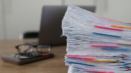 Detailansicht eines Aktenstapels, im Hintergrund verschwommen Taschenrechner mit Brille daraufliegend