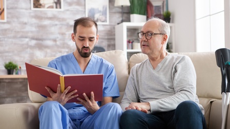 Zwei Personen sitzen nebeneinander auf einem hellen Sofa. Die linke Person trägt einen blauen Kittel und liest aus einem Buch vor. Die rechte Person hat graue Haare und trägt eine Brille. Neben ihr sind Krücken.