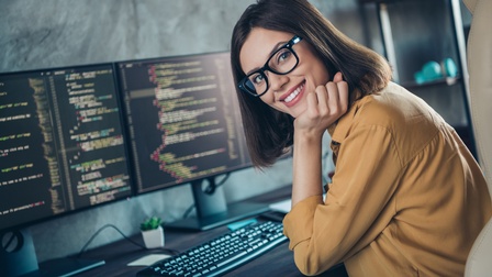 Lächelnde Person mit Brillen stützt Kopf in die Hand an Schreibtisch mit Monitoren, auf denen Quellcodes zu sehen sind, sitzend