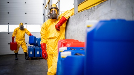 Person in gelber Schutzkleidung, mit roten Handschuhen, und Atemmaske lehnt an Mauer, im Vordergrund blaue und rote Kanister, im Hintergrund weitere Person in selber Schutzkleidung beim Schlichten von Kanistern