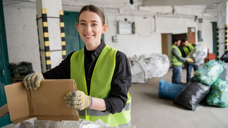 Lächelnde Person in gelber Warnweste mit Handschuhen faltet Papierkartonage, im Hintergrund verschwommen weitere Personen gefüllte Müllsäcke inspizierend