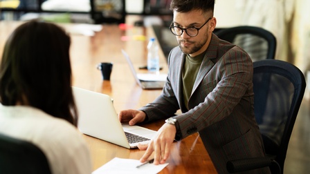An einem Tisch sitzen zwei Personen. Die rechte Person schiebt der anderen Person einen Zettel und einen Stift hin