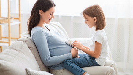 Kind sitzt bei einer schwangeren Person auf der Couch und hält Babyschuhe auf den Bauch