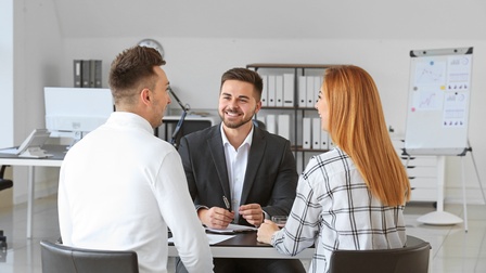 Drei Personen an Tisch in weißgrauem Büroraum sitzend im Gespräch miteinander: Zwei Personen in Rückenansicht, eine Person in Frontalansicht lächelnd, im Hintergrund verschwommen Regale mit Ordnern, Tisch mit Computer und Flipchart