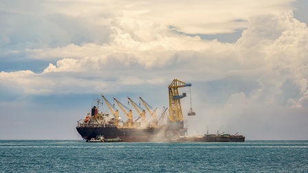 Mehrere Frachtschiffe auf dem offenen Meer. Neben ihnen steht ein größeres Schiff, auf dem vier Kräne sind