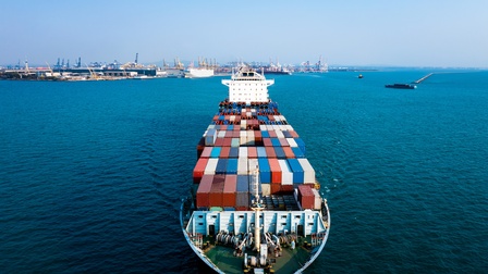 Ein Frachtschiff mit vielen Containern, das auf einem Gewässer fährt. Im Hintergrund ist ein Hafen mit vielen Hafenkränen