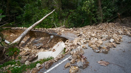 Zerstörter Straßenabschnitt in Kurve verlaufend voller Steingeröll, ein entwurzelter umgekippter Baum liegt über der Straße, im Hintergrund grüne Sträucher und Bäume