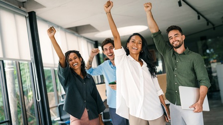 Porträt von vier lächelnden Personen in Bürokleidung, die nebeneinander in einem Büro stehen und jeweils die rechte Faust in die Luft gestreckt haben. Eine Person hält einen Laptop, eine Person ein Tablet, eine Person ein Handy
