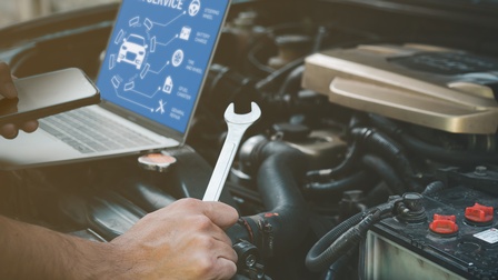 Eine Person steht an einer geöffneten Motorhaube. Die Person hält einen Drehschlüssel und ein Handy. Am Rand der geöffneten Motorhaube steht ein aufgeklappter Laptop, auf dem Autoservice steht