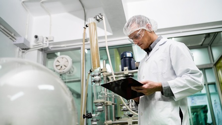 Person in weißem Kittel, mit Haarnetz und Schutzbrille steht in Laborraum mit labortechnischen Apparaturen und blickt auf Clipboard, das sie in Händen hält