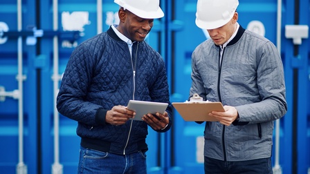 Frontalaufnahme zweier nebeneinander stehenden Personen mit weißen Helmen und Jacke. Die linke Person hält ein Tablet, die rechte ein Klemmbrett. Hinter den Personen sind blaue Container